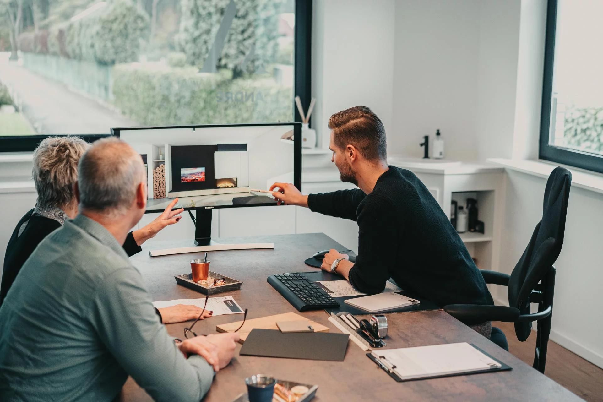 Een ontwerper bespreekt een interieurconcept met klanten aan een bureau, met focus op een 3D-model op een computerscherm.